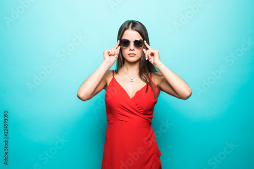 Elegant young attractive woman wearing red summer dress with hands on sunglasses isolated over pastel blue background.