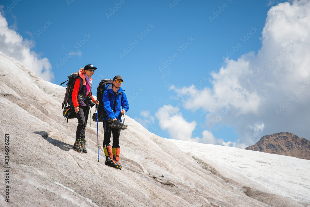 Two tourists, a man and a woman with backpacks and cats on their feet, stand on the ice in the background of the mountains of the sky and clouds. Watch in the distance