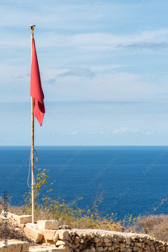 Fototapeta premium Red flag on a pole. Clouds and sea view background. Vertical. No wind