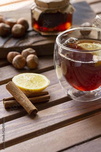 Tea in glass cup with lemon and vanilla pod at wood background. teapot with tea near. Huzelnut and jam in jar at wood board behind. photo