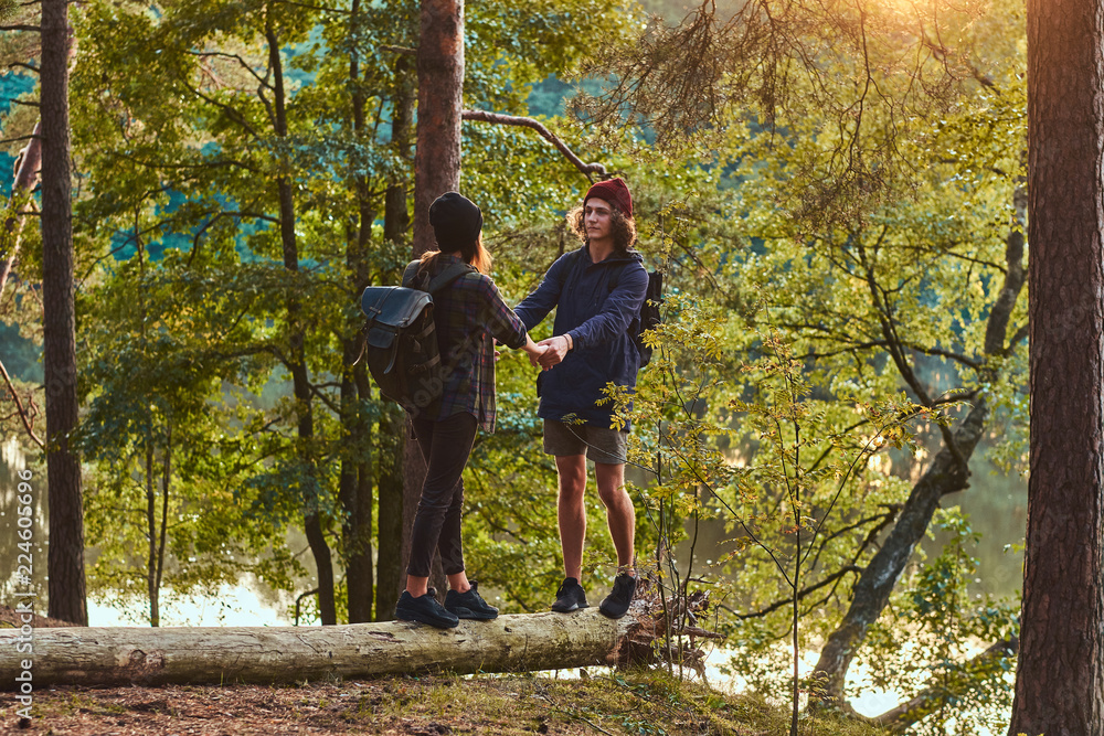 Fototapeta premium Young hipster couple holding hands standing on a tree trunk in a beautiful forest at sunset. Travel, tourism, and hike concept.