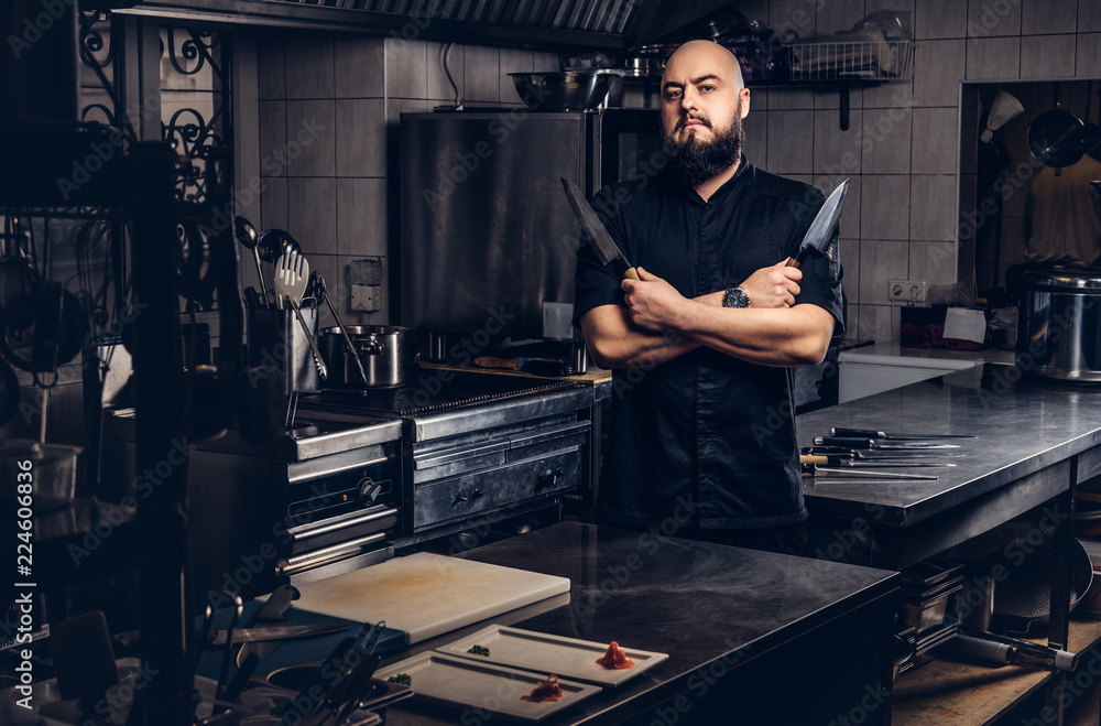 Obraz premium Bearded cook in black uniform holds knife and standing with crossed arms in the kitchen.
