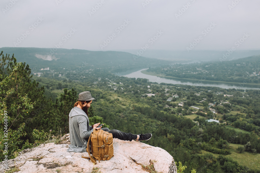 Young tourist with a beard resting on the High Mountain and responding to the message of friends on the Internet.Young man dressed in stylish clothes on the head hat behind the backpack