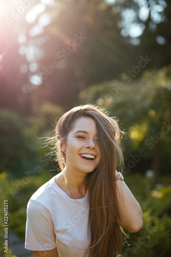 Positive human emotions. Close-up portrait of a young cheerful girl with long blonde hair on a blurred green background. The model in a white t-shirt openly smiles, laughs and looks at the camera.