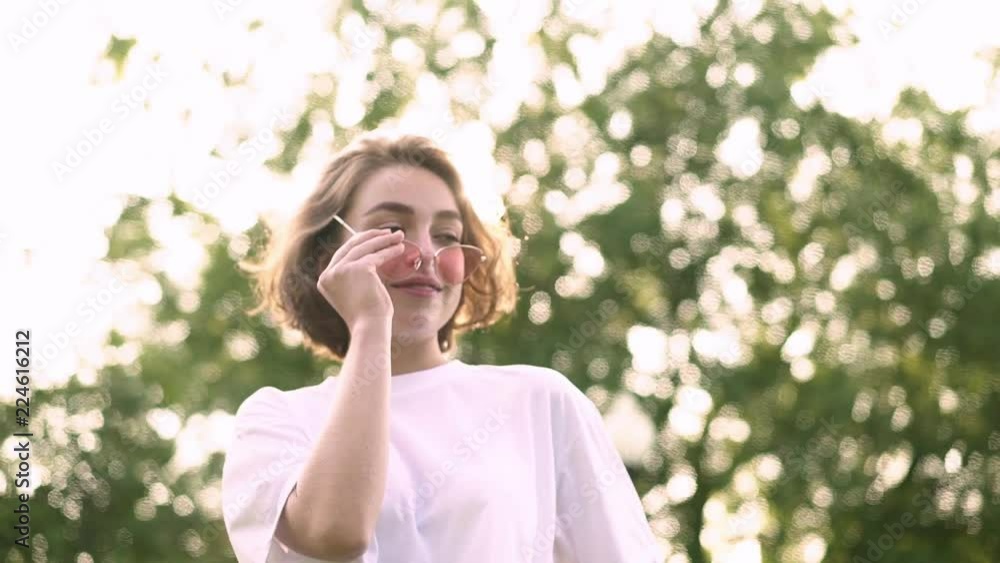 Cute young woman in white t shirt walking in summer park, playing with her short fair hair and putting on pink sunglasses. Concept of fashion and happiness. Tracking slow motion low angle shot