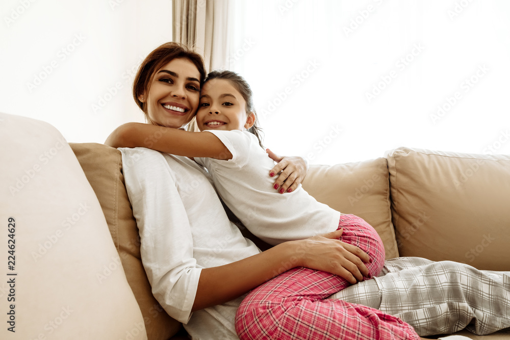 Family. Love. Mom and daughter in pajamas are hugging, looking at camera and smiling while sitting on couch at home