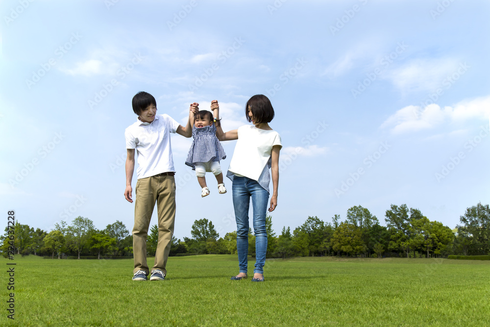 青空をバックに芝生の公園で幼い女の子と手を繋ぐ若い夫婦。幸せ、家族、親子、愛、絆イメージ