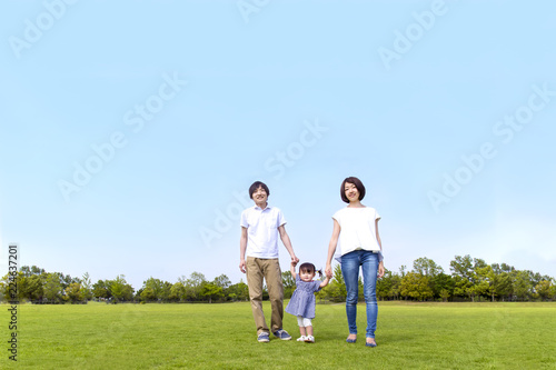 青空をバックに芝生の公園で幼い女の子と手を繋ぐ若い夫婦。幸せ、家族、親子、愛、絆イメージ