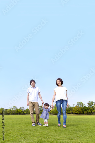 青空をバックに芝生の公園で幼い女の子と手を繋ぐ若い夫婦。幸せ、家族、親子、愛、絆イメージ