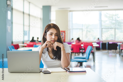 young asian female college student smiling at camera while sitting in library with laptop.