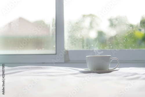 Fresh morning coffee on the white bed near window in the sunny day, green nature background. Lifestyle Concept