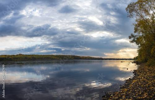 Panoramic image of a calm river with blue waters  on the banks of which trees with autumn coloring rise  the sky is strewn with clouds through which a ray of light tries to penetrate.