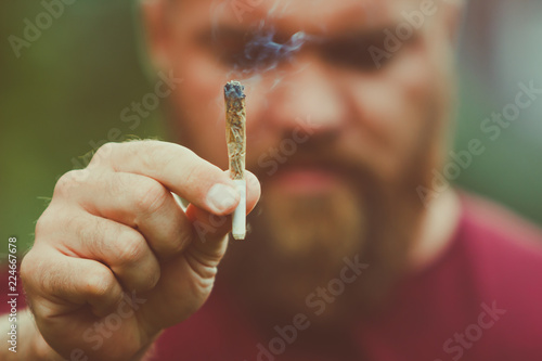 Sad or disappointed young man with beard is holding a smoking marijuana joint - legalization (or legalisation) medical cannabis concept - smoke is coming out of the weed or hashish cigarette