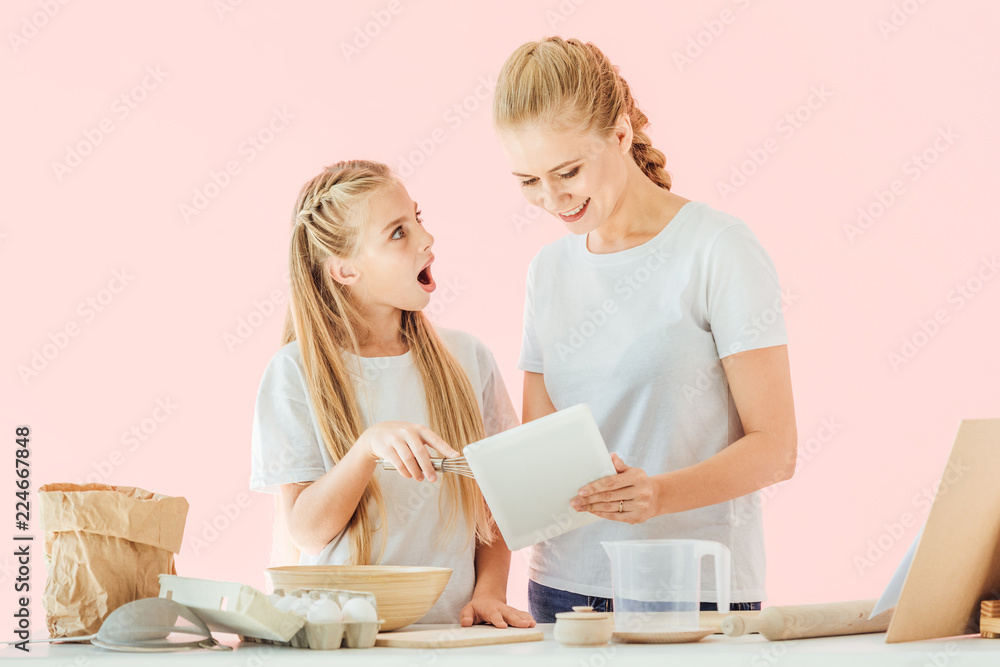 happy mother and surprised daughter in white t-shirts using tablet while cooking isolated on pink happy mother and surprised daughter in white t-shirts using tablet while cooking isolated on pink
