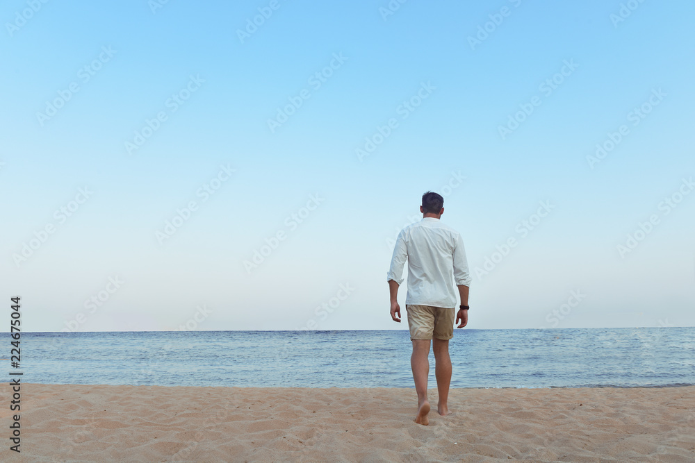 Handsome man in white shirt standing on a beach looking hot on summer day enjoying life. Brutal Male model poses with wet short in water naked chest out, smiling with smirk on his look and kind eyes.