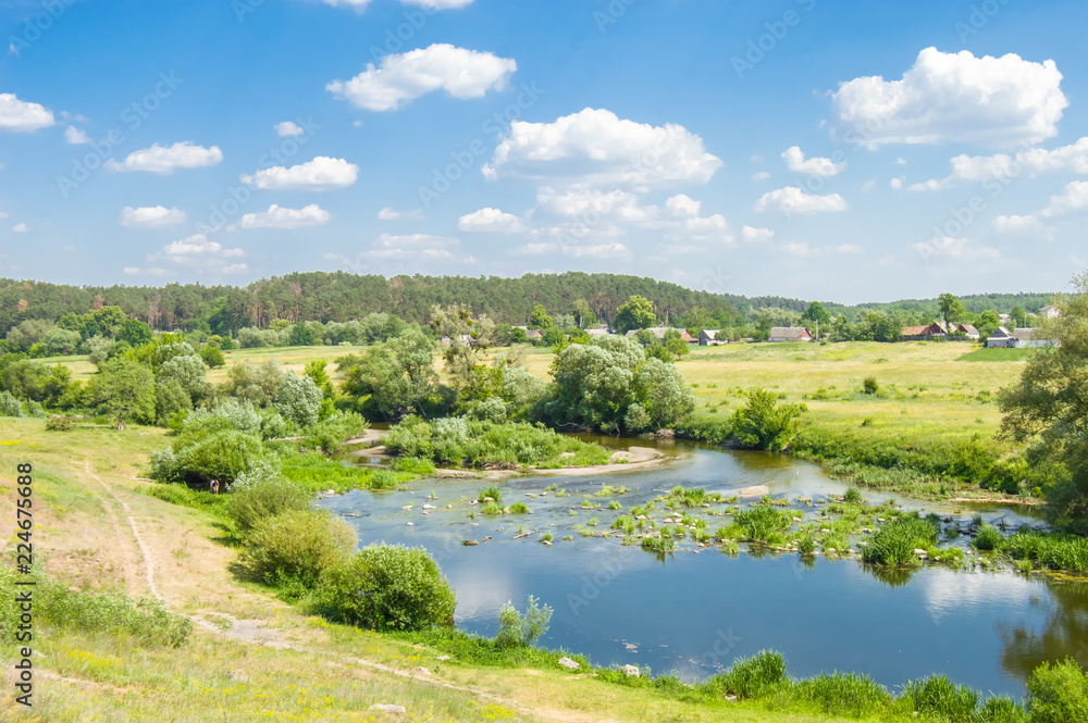 The River Teteriv (Teterev). Stone obstacles - rapids or rifts on the river bed amid a forest with blue skies, rural landscape with houses away on background and water meadows of the river