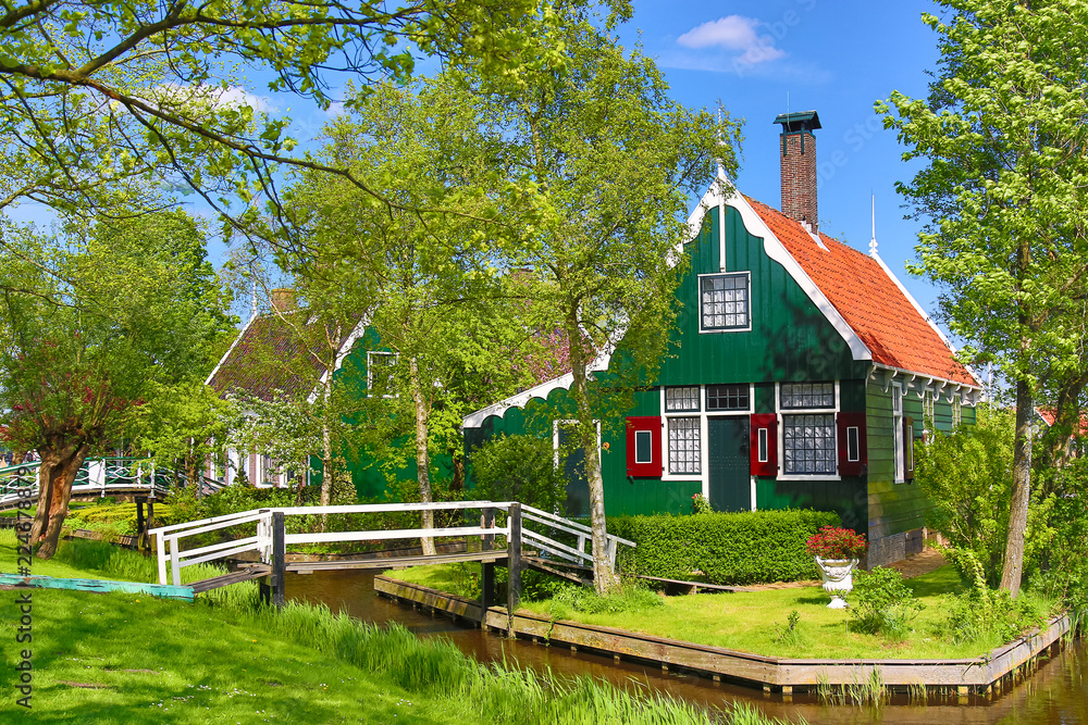 Naklejka premium Tradycyjny zielony holenderski dom z drewnianym mostkiem na tle błękitnego nieba w wiosce Zaanse Schans, Holandia. Znane miejsce turystyczne
