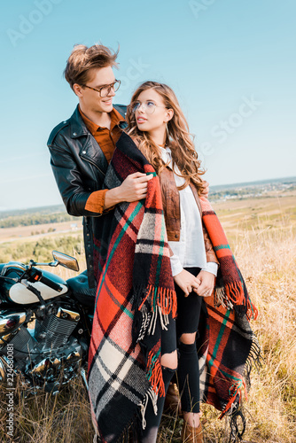 young couple in glasses standing near vintage motorbike and looking at each other