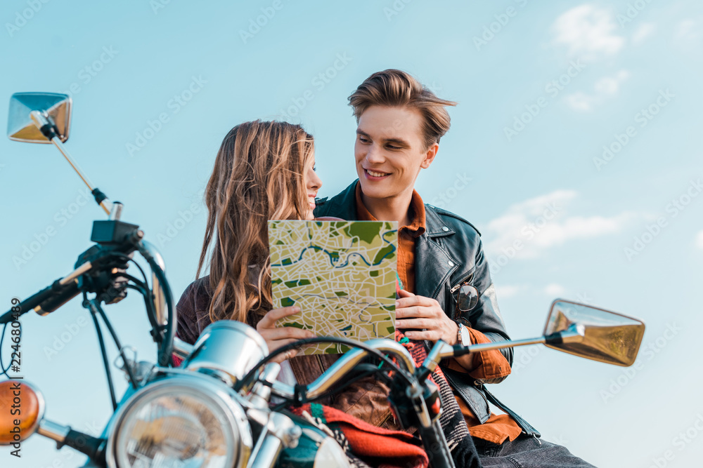 young couple of travelers holding map and sitting on motorbike against blue sky