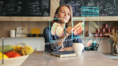 Blonde pretty woman in the kitchen uses transparent hologram futuristic screen to purchase vegetables online, future shopping concept