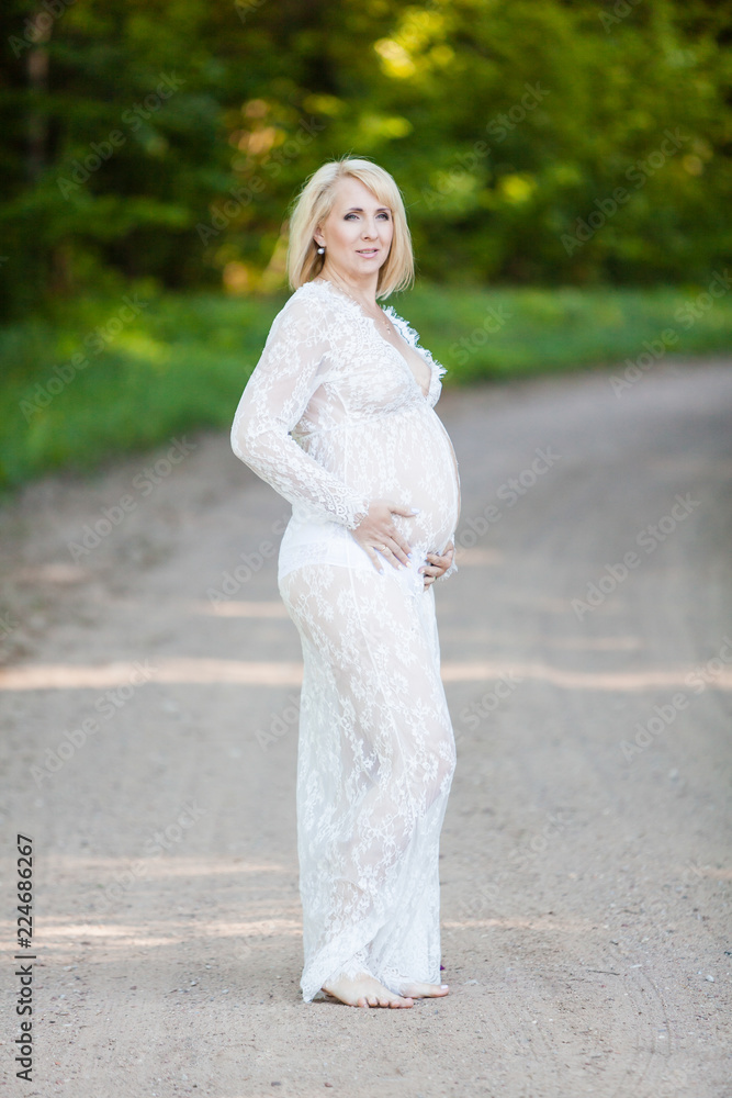 Beautiful pregnant woman in sheer long lacy white maternity dress looking dreamy on lonely road in forest Beautiful pregnant woman in sheer long lacy white maternity dress looking dreamy on lonely road in forest