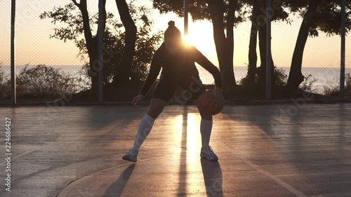 Young caucasian female basketball player dribbling and practicing ball handling skill on court. Morning dusk, sun shines on the background photo