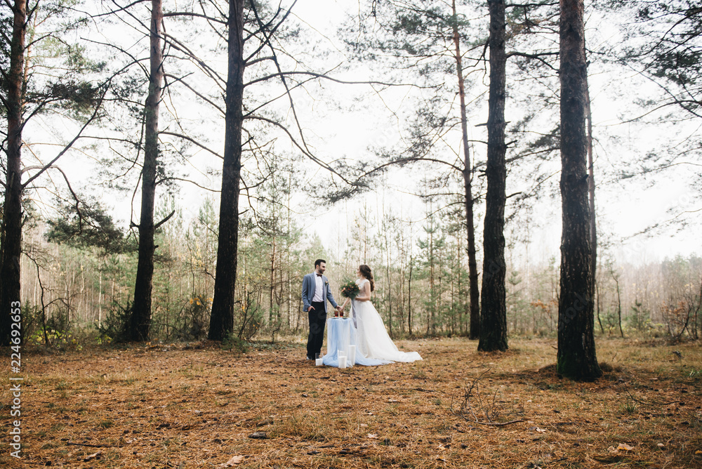 Happy bride and groom stand near the table, set for two in the woods. Autumn. The concept of a romantic date Happy bride and groom stand near the table, set for two in the woods. Autumn. The concept of a romantic date