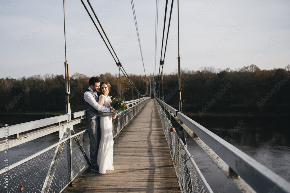 Happy young smiling bride and groom are standing on the suspension bridge. Wedding photos in an interesting place