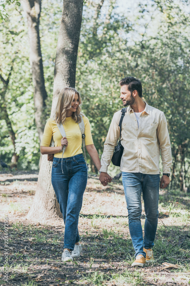 smiling young couple with backpacks holding hands while walking in park smiling young couple with backpacks holding hands while walking in park