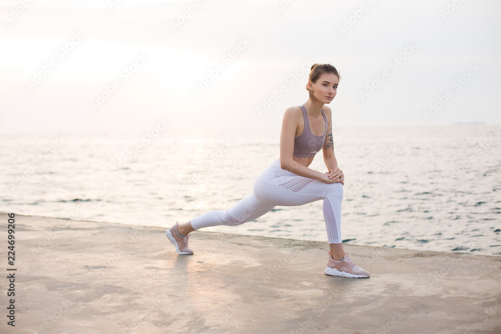 Thoughtful girl in sporty top and white leggings practicing yoga with amazing sea view on background. Young woman dreamily looking in camera while training by the sea Thoughtful girl in sporty top and white leggings practicing yoga with amazing sea view on background. Young woman dreamily looking in camera while training by the sea