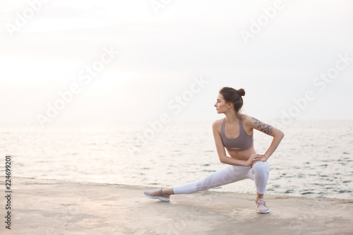 Thoughtful girl in sporty top and leggings practicing yoga with amazing sea view on background. Young woman stretching by the sea