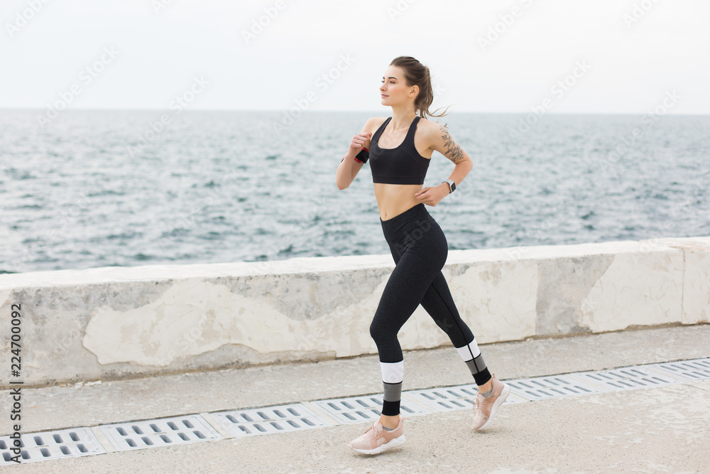 Young beautiful woman in dark gray sporty top and leggings dreamily looking aside while running with sea view on background