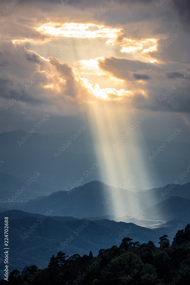 Bright sunlight shining through hole of clouds to dark scene of mountain range before sunset in Thailand rainforest area.