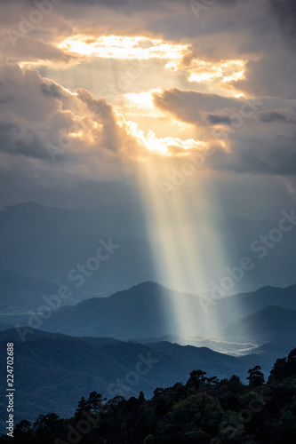 Bright sunlight shining through hole of clouds to dark scene of mountain range before sunset in Thailand rainforest area.