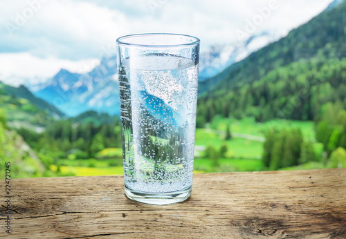 Glass of water on the stone. Blurred snow mountains tops and green forests at the background.