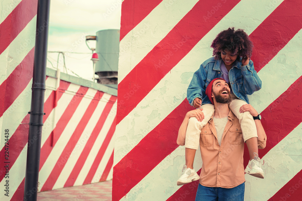 Looking up. Emotional bearded young man putting his head up and smiling to his beloved woman while carrying her on his shoulders