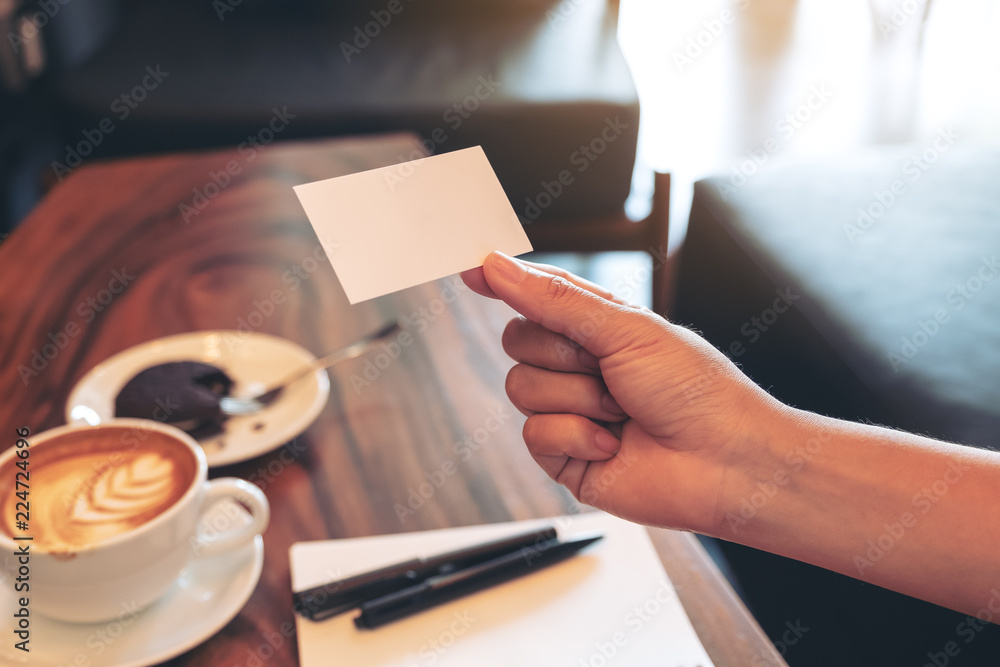 Obraz premium A hand holding white empty business card with notebook and coffee cup on wooden table
