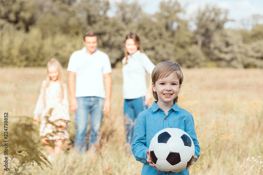 happy little kid holding ball while his family standing blurred on background in field happy little kid holding ball while his family standing blurred on background in field