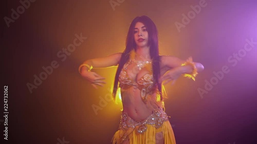 Alluring sexy traditional oriental belly dancer girl dancing on yellow neon smoke background. Woman in exotic costume with feathers sexually moves her semi-nude body