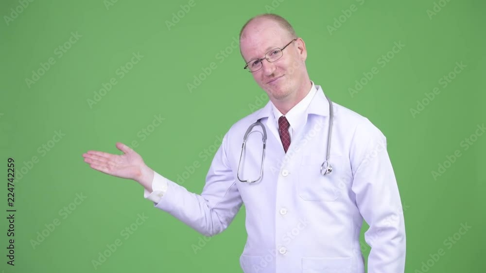 Happy mature bald man doctor showing something against green background Happy mature bald man doctor showing something against green background