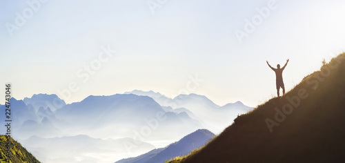 Man on peak of mountain. Emotional scene. Young man with backpack standing with raised hands on top of a mountain and enjoying mountain view. Hiker on the mountain top. Sport and active life concept.
