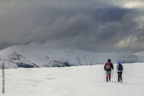 Winter mountain landscape. Back view of travelers tourist hikers with backpacks on snowy field walking towards distant mountain on cloudy dark blue stormy sky copy space background on cold winter day.