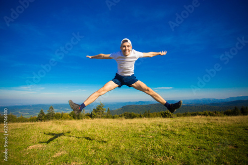 Summer hiking in mountains. Young tourist man in cap with hands up on top of mountains admires nature. Travel concept