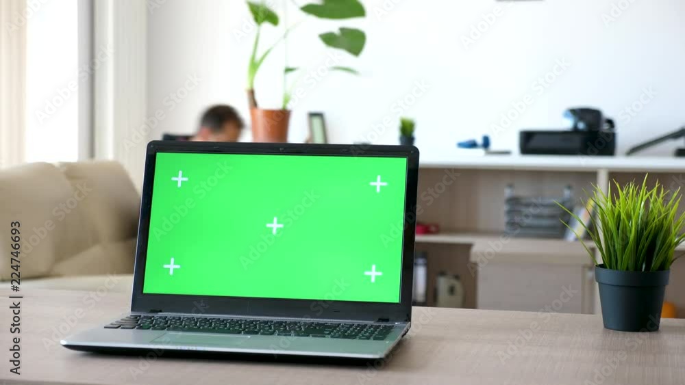 Laptop on a desk in the living room with a isolated green screen and marks for tracking. Man walks in the room and sits on the couch in the background Laptop on a desk in the living room with a isolated green screen and marks for tracking. Man walks in the room and sits on the couch in the background