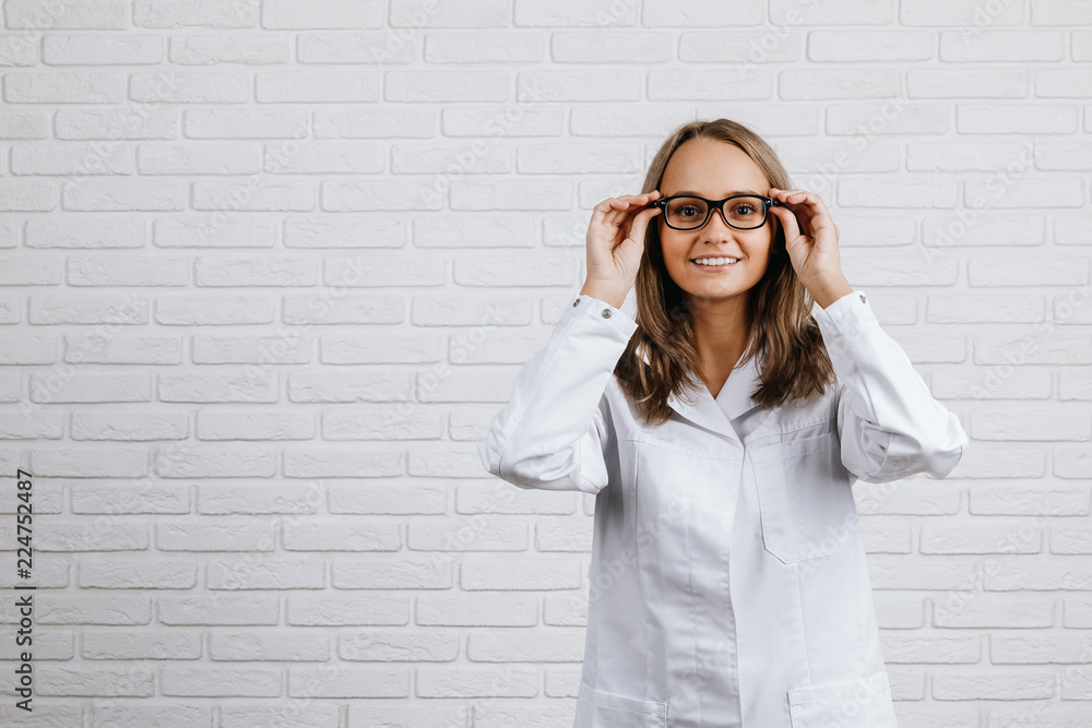 Εκτύπωση καμβά The girl doctor in a white coat put on black glasses   and smiling on white back