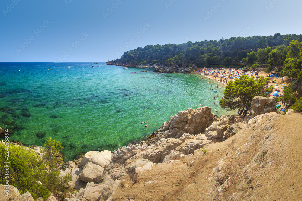 View on Treumal beach in Lloret de Mar, Costa Brava, Spain on sunny summer day. Cala Treumal beach. Vacation on tropical mediterranean sea. Turquoise water, rocks, green plants, clear sky.