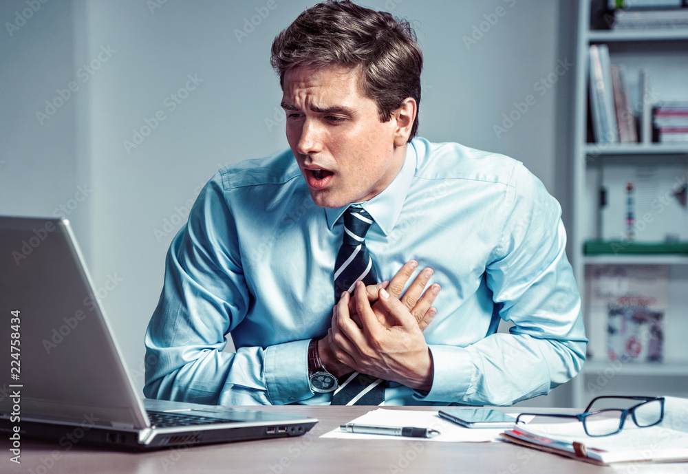 Naklejka premium Office manager having heart attack. Photo of young man holding hand on painful chest in the workplace. Medical concept.