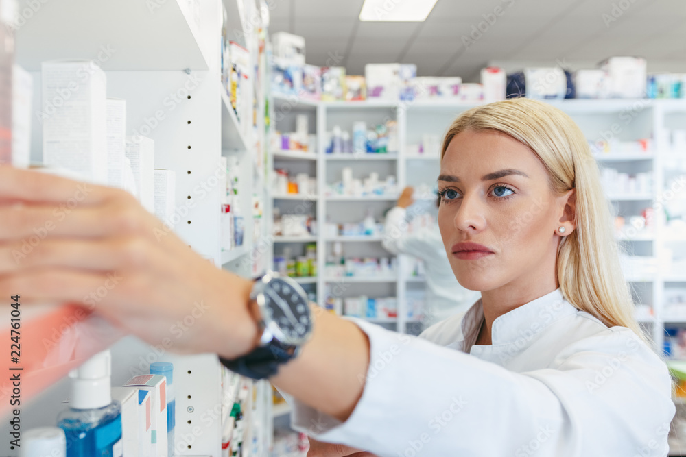 Serious woman pharmacist reaching for a medications among shelves at pharmacy drugstore