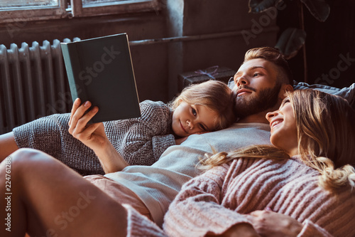Mom, dad and daughter reading storybook together while lying on bed. photo