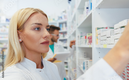 Serious woman pharmacist reaching for a medications among shelves at pharmacy drugstore